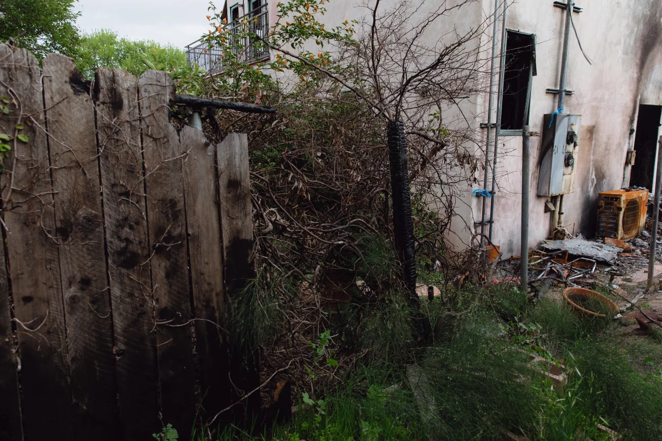 Fire damage to residential exterior: scorched siding, destroyed fence line, and burnt vegetation