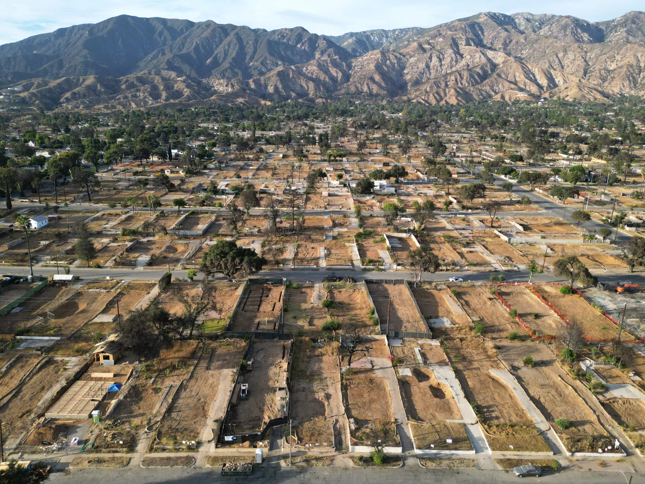 Aerial view of cleared residential lots and foundations after a wildfire or disaster, with rugged mountains in the background