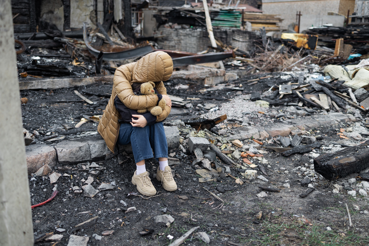 A distressed child, wearing a tan hooded puffer coat, sits clutching a teddy bear amidst the charred rubble and debris of a bombed or burned-down building