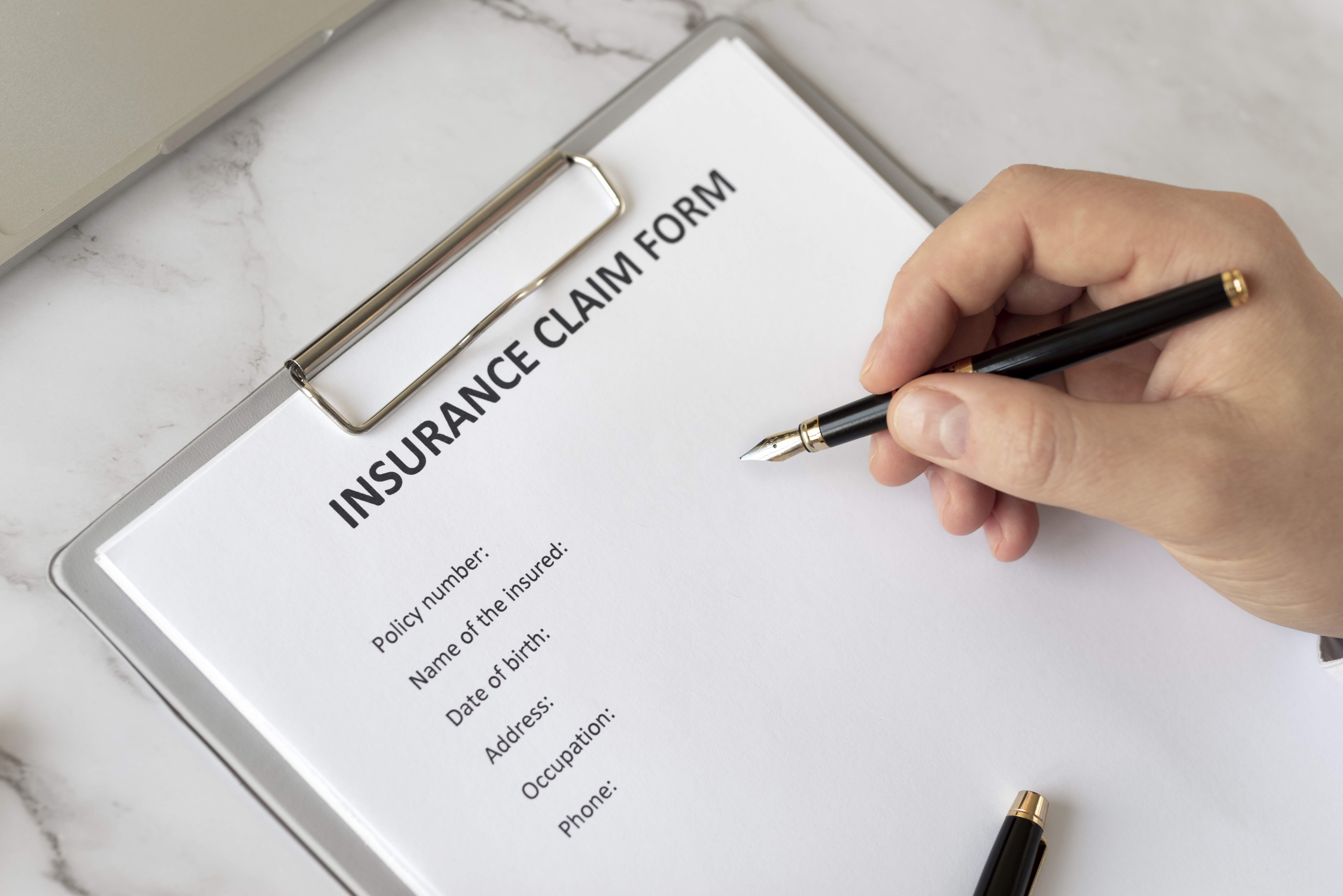 Close-up view of a hand holding a pen and preparing to fill out a general Insurance Claim Form on a clipboard with a marble background