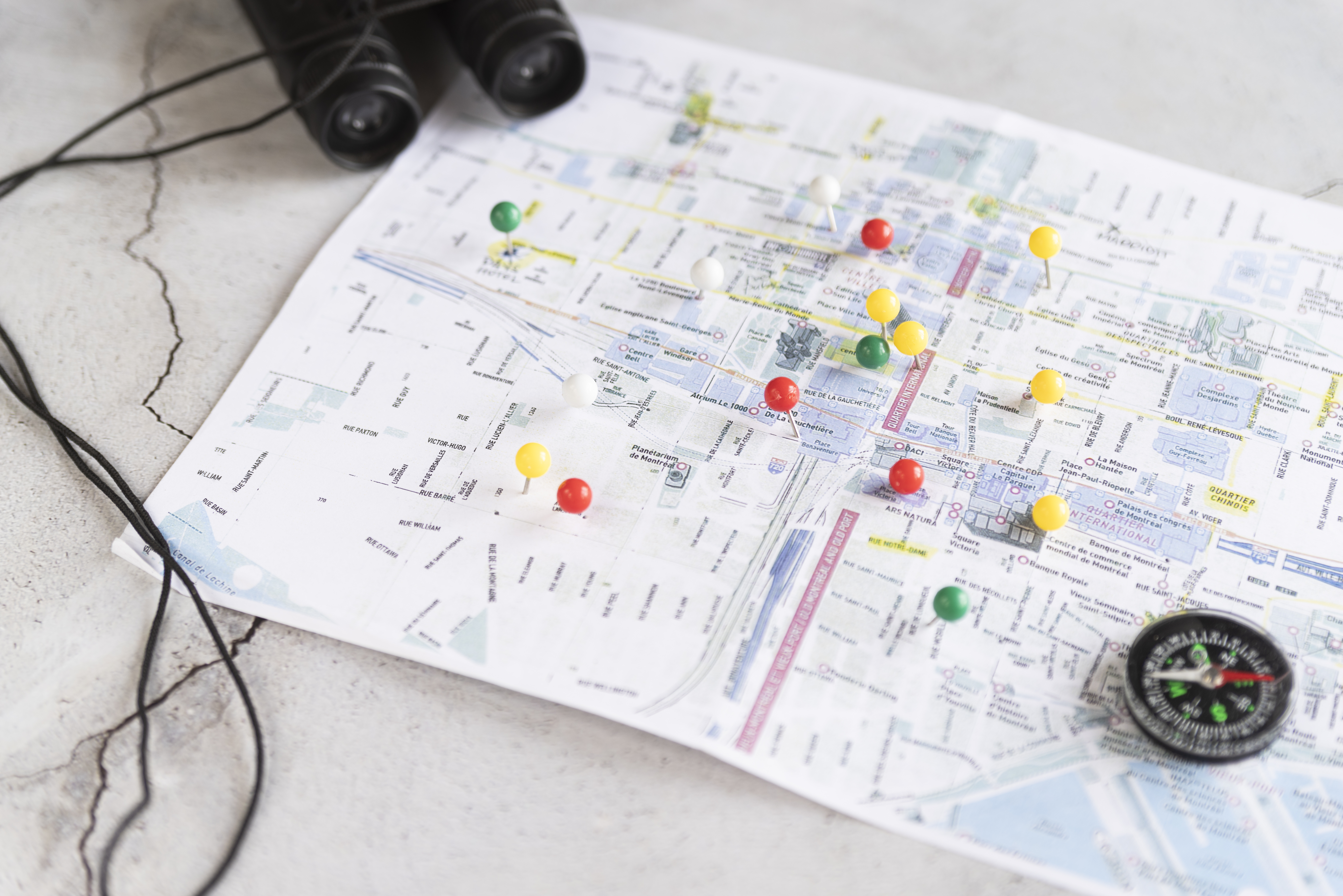 Overhead view of a paper city map marked with multiple colored pushpins, next to a pair of binoculars and a small magnetic compass, representing travel planning or location targeting