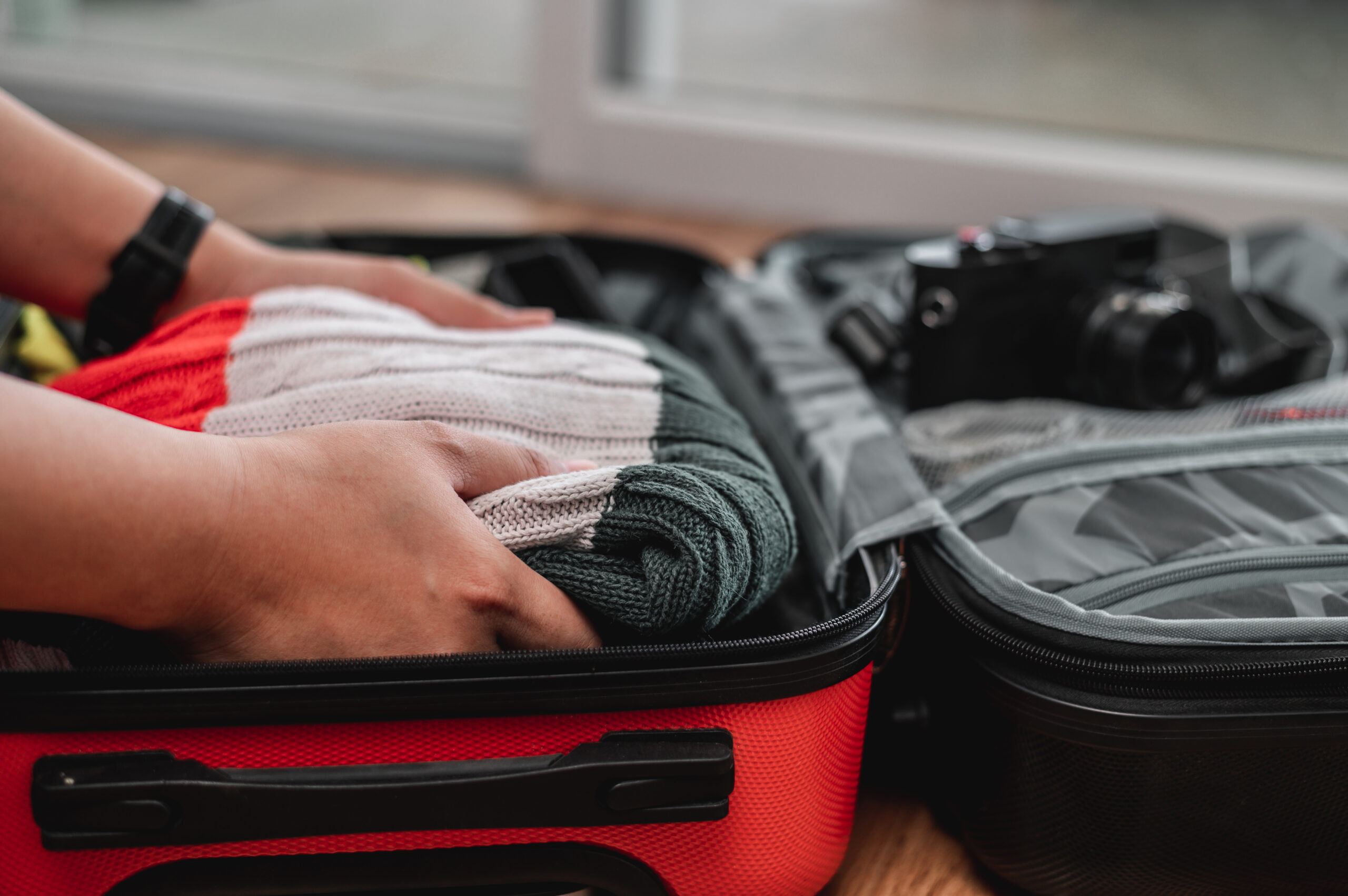 Close-up of hands folding and packing a knitted sweater and other clothes into an open red suitcase on a wooden floor, with a black vintage camera visible nearby