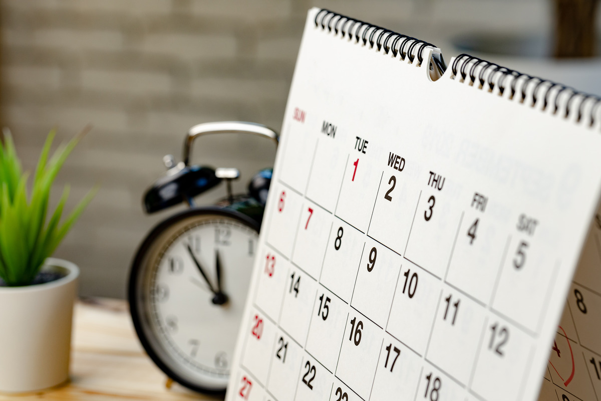 Desk calendar showing monthly schedule with dates marked, next to alarm clock and plant, representing time management, planning, and organization