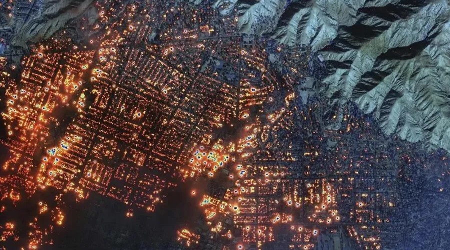 Aerial view of wildfire spreading across urban area at night, showing city grid with active fire hotspots and surrounding mountainous terrain