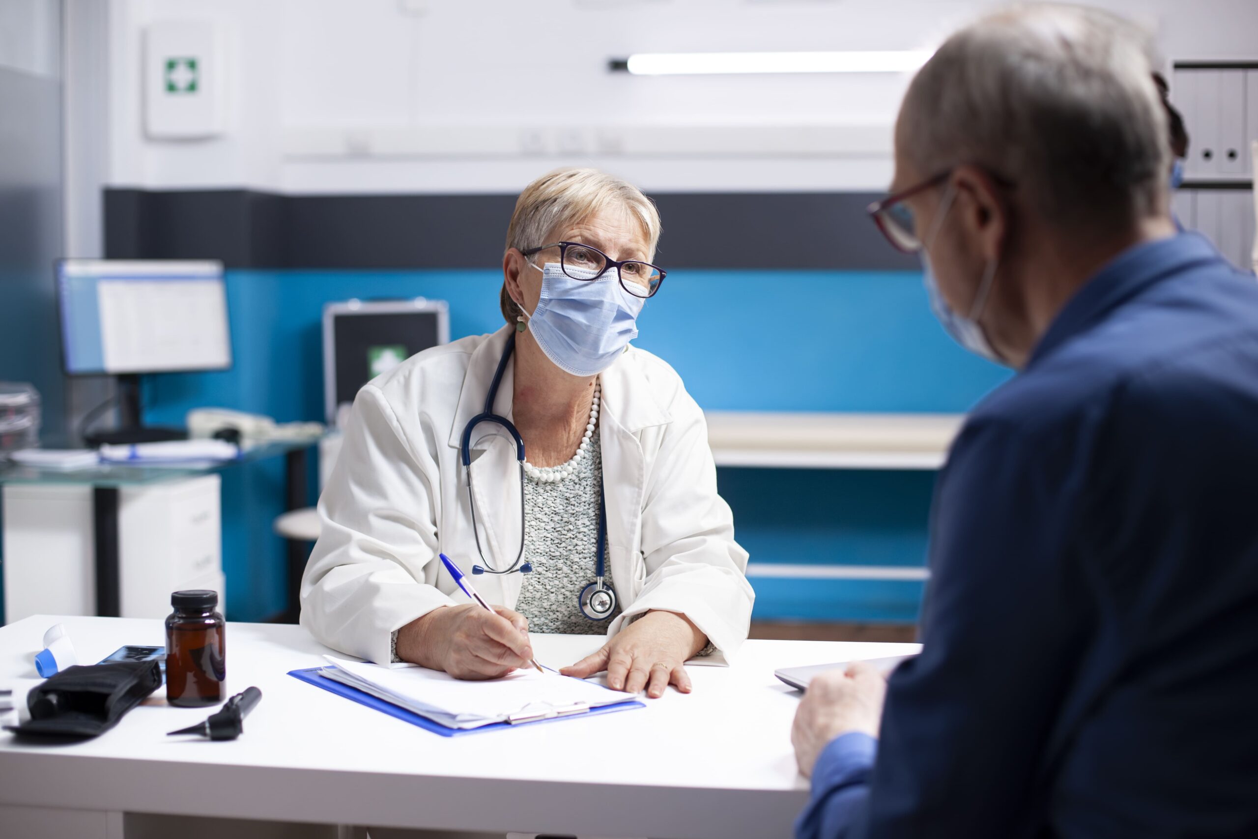 Doctor consulting with an older patient in a medical office, both wearing face masks, representing healthcare consultation, patient care, medical evaluation, treatment planning, and physician-patient communication.