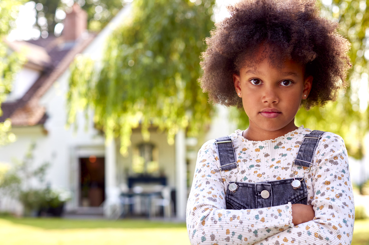 Young child standing outdoors in front of a home with arms crossed and a serious expression, representing child welfare, family dynamics, emotional well-being, childhood resilience, and family-focused support services.